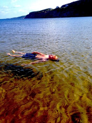 A person lying on a beach with clear water, a rocky shoreline, and a forested hill in the background. - Year-round Vacation Rental - Fly Fishing, Golf, Beaches, Cabot Trail & More.
 (Margaree)