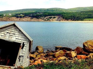 A wooden boathouse by a rocky shoreline with a calm lake and forested hills in the background. - Year-round Vacation Rental - Fly Fishing, Golf, Beaches, Cabot Trail & More.
 (Margaree)