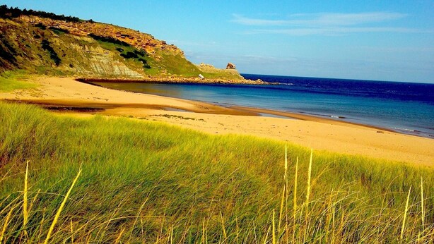 Un paysage côtier avec une plage de sable, un premier plan herbeux et une falaise rocheuse sur laquelle se trouve une petite construction.