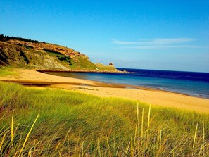A coastal landscape with a sandy beach, grassy foreground, and a rocky cliff with a small structure. - Year-round Vacation Rental - Fly Fishing, Golf, Beaches, Cabot Trail & More.
 (Margaree)