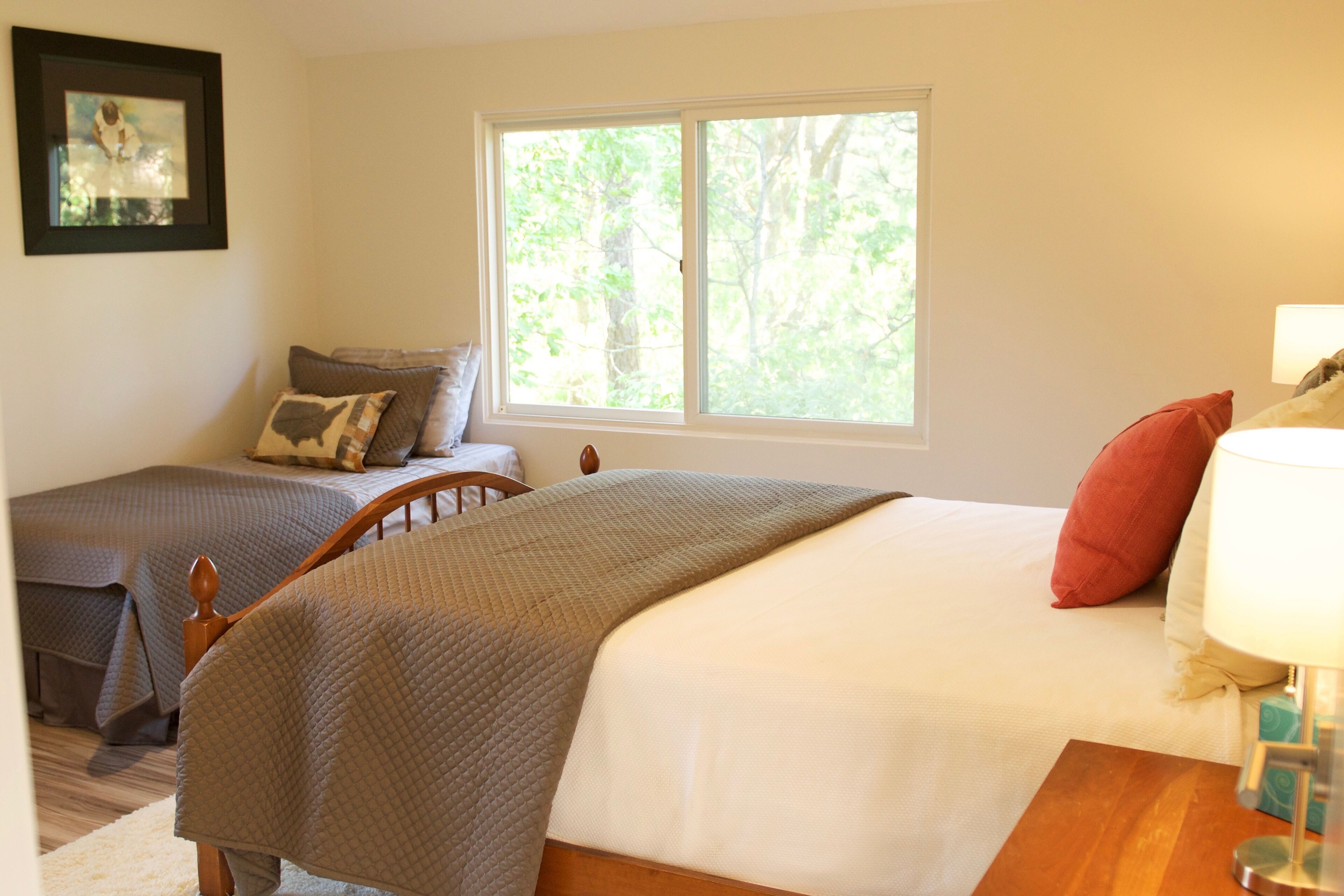 A bedroom with two beds, a window with a view of trees, a framed picture on the wall, and a bedside table with a lamp.