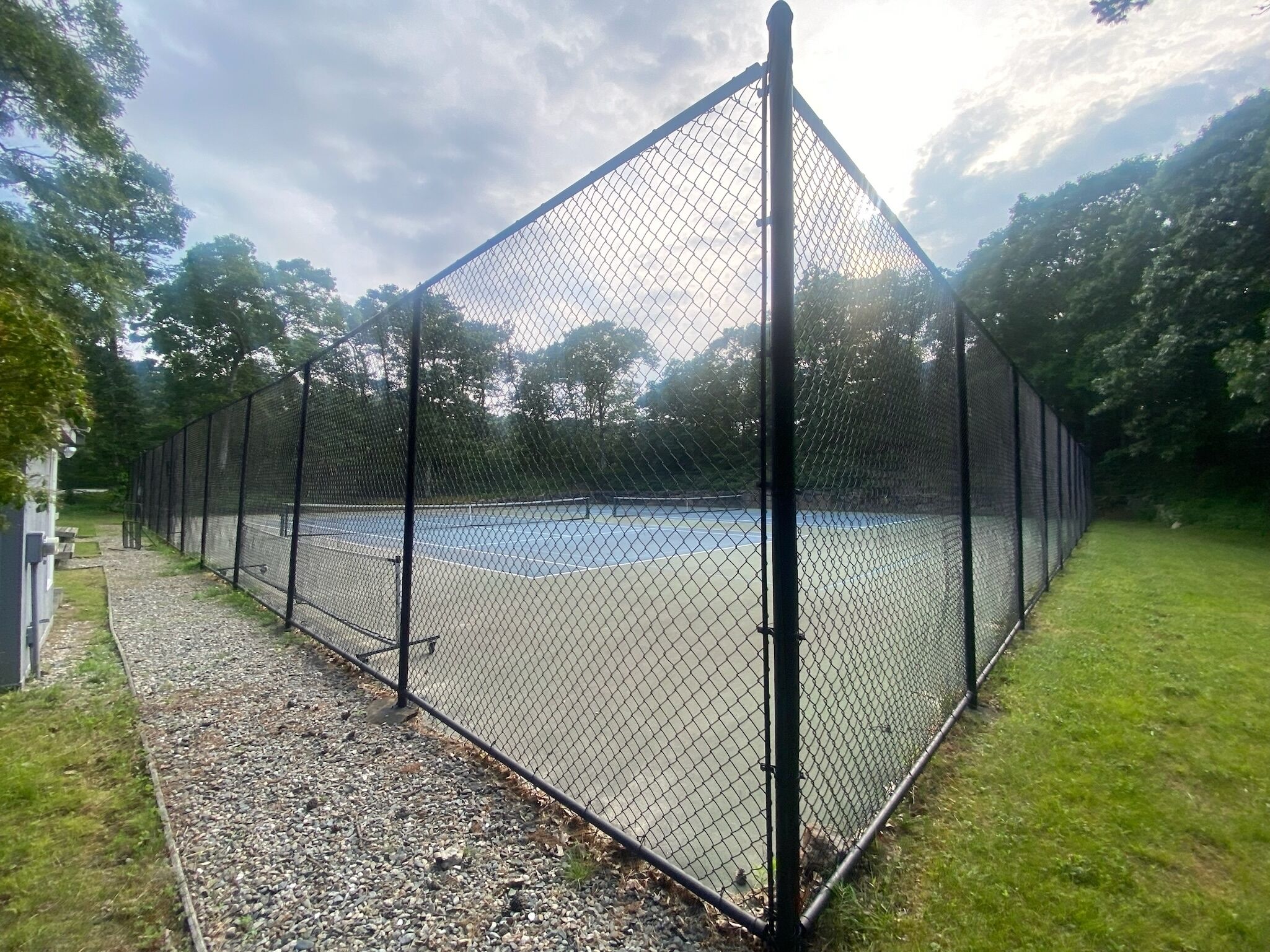 A tennis court enclosed by a chain-link fence with a gravel path leading to it.