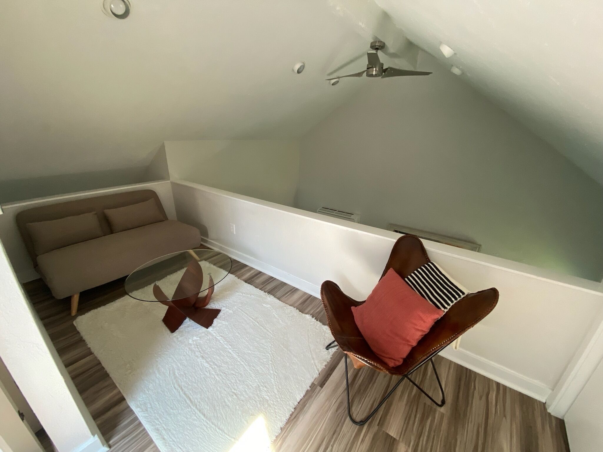 Ein Loft-Wohnraum mit hoher Decke, Deckenventilator, braunem Ledersessel mit rotem Kissen, beigem Sofa und weißem Teppich.
