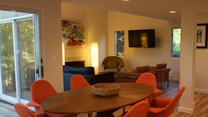 Un salon moderne avec une table à manger en bois, entourée de chaises orange, d’un canapé bleu et d’un téléviseur fixé au mur.
