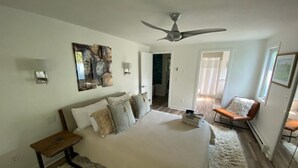 Une chambre avec un grand lit, un ventilateur de plafond, un banc et une chaise près de la fenêtre.