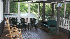 Une véranda couverte avec un plancher en bois, des chaises vertes, un fauteuil à bascule et un banc.