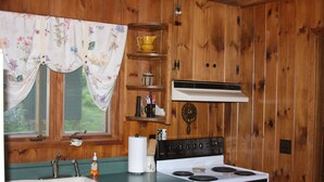 Une cuisine aux murs en bois, avec un poêle blanc, un évier et une fenêtre ornée de rideaux à motifs floraux.