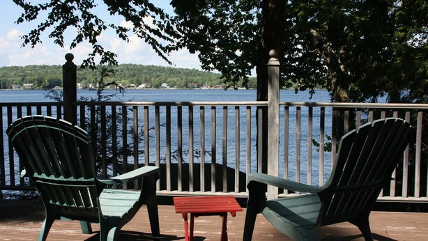 Une terrasse en bois avec deux fauteuils Adirondack et une petite table rouge, donnant sur une étendue d’eau dont on aperçoit la rive au loin.