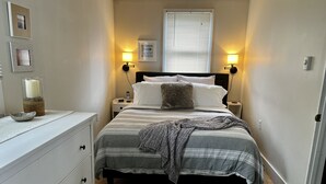 Una camera da letto con un letto, comodini, una cassettiera, una finestra con persiane, luci a parete e un quadro incorniciato.