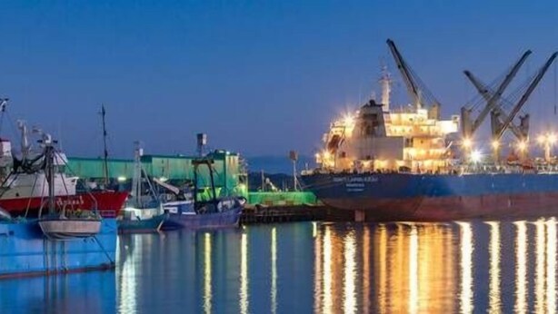 A large cargo ship docked at a port with cranes and other vessels nearby.