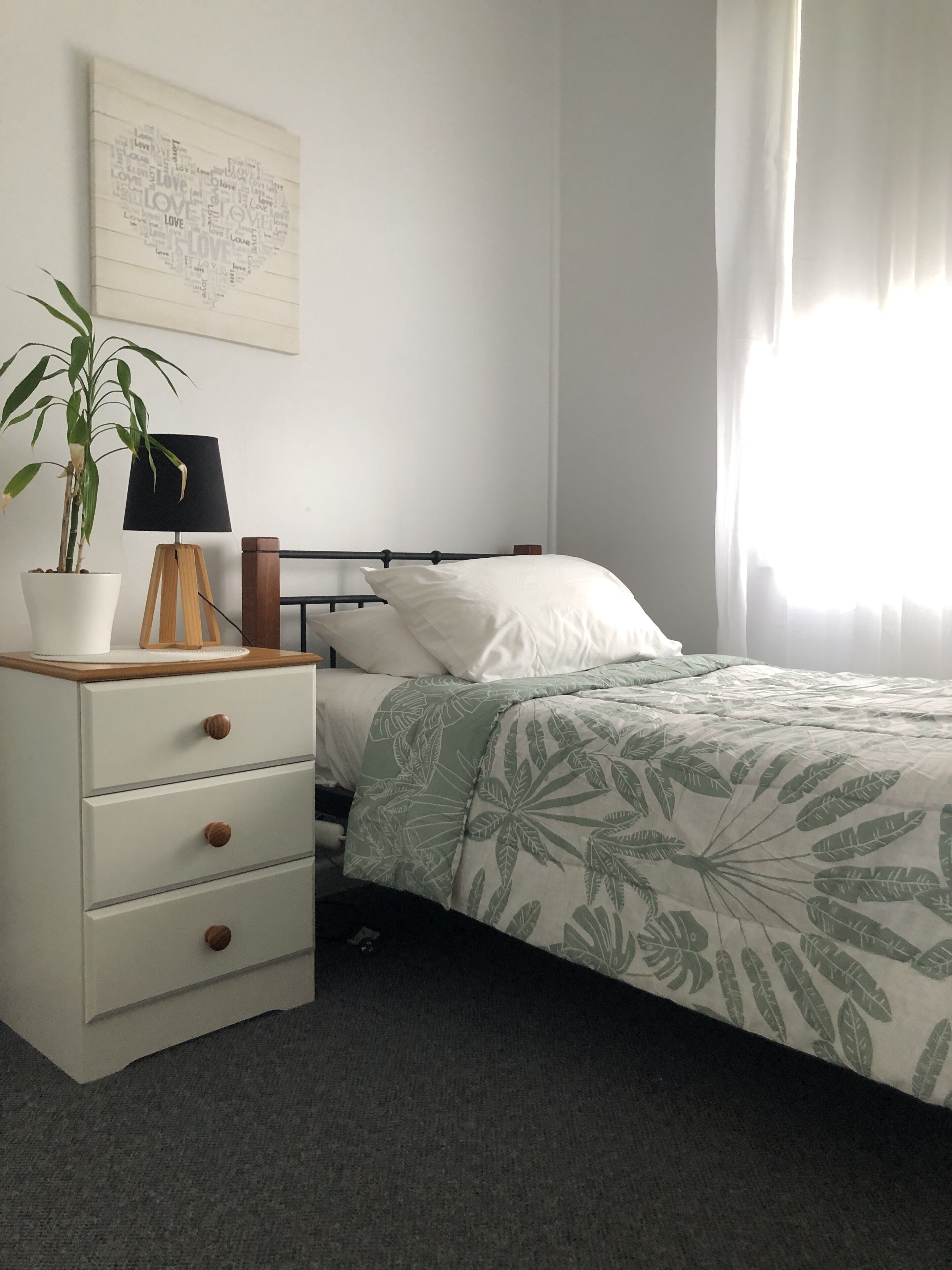 Une chambre à coucher comprenant un lit, une table de chevet, une lampe, une plante en pot et une œuvre d’art murale.