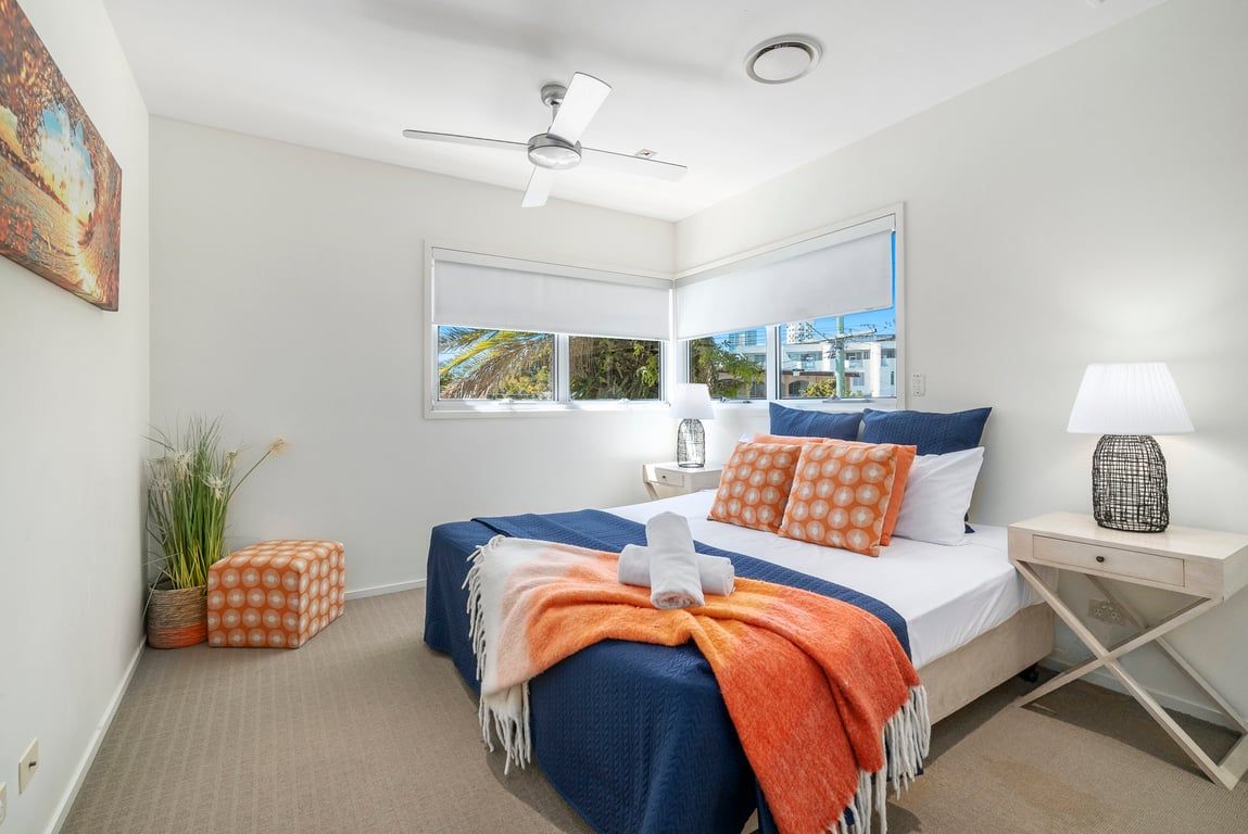 A hotel room with a bed, a ceiling fan, a window with a view, a bedside table with a lamp, and a decorative plant.