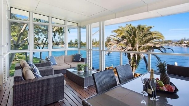 A sunlit outdoor lounge area with wicker furniture, a glass table, and a view of a pool and palm trees. - Magical Water Views for the whole family (Gold Coast)