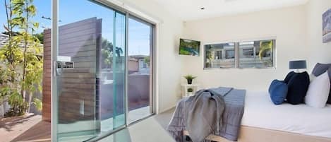 Une chambre moderne dotée d’une grande porte-fenêtre coulissante donnant sur un balcon, d’un lit avec de nombreux oreillers, d’une petite table de chevet avec une lampe et d’un téléviseur fixé au mur.
