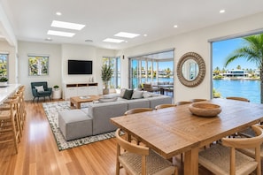 A modern living room with a large TV, a dining area with a wooden table and chairs, and a view of a body of water through large windows.