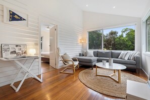 A modern living room with a grey sectional sofa, a round coffee table, and a white armchair. There is a large window providing a view of trees and a whiteboard with a map and text. - Fish at Freycinet - Freycinet Holiday Houses (Coles Bay)