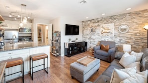 A modern kitchen with a central island, a living room with a grey sofa and ottoman, and a stone accent wall. - Peaceful Sports Village Condo | King Bed, Balcony & Pools (St. George)