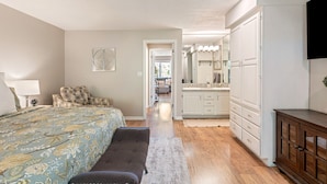 A bedroom with a bed, bench, and a hallway leading to a bathroom with a sink and mirror. - Peaceful Sports Village Condo | King Bed, Balcony & Pools (St. George)