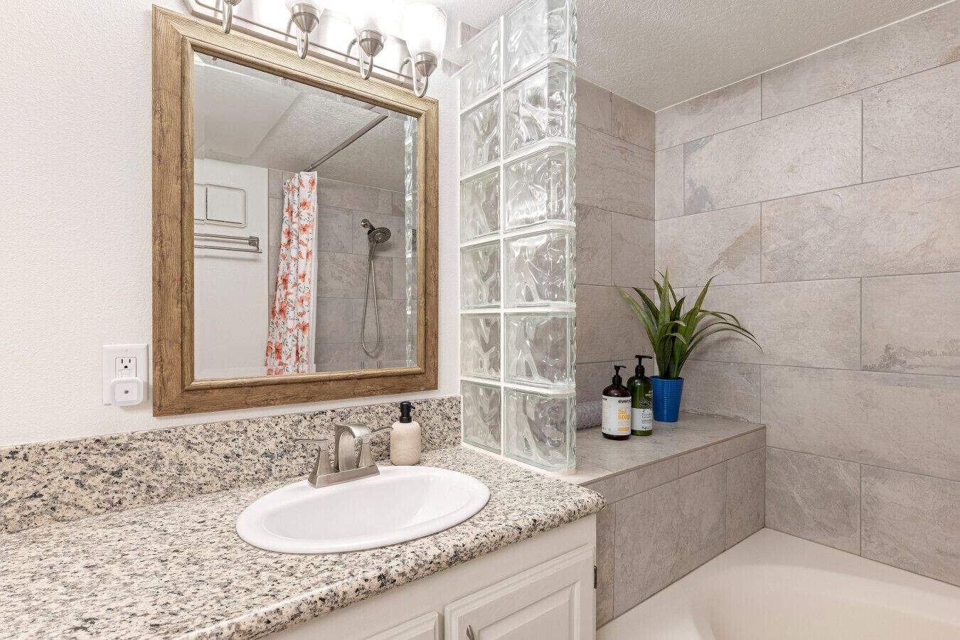 Une salle de bain avec un comptoir en granit, un lavabo blanc, un miroir avec un cadre en bois, une douche avec une paroi en verre et une plante en pot.