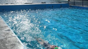 Ein Schwimmbad mit klarem, blauem Wasser, eine Person schwimmt darin, und im Hintergrund Meer und Klippen.