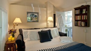 A hotel room with a bed, two bedside lamps, a painting, a bookshelf, and a bathroom visible through an open door.