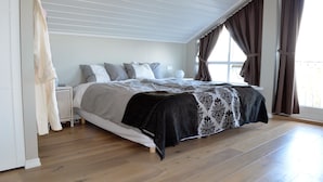 A bedroom with a large bed, a bedside table, a window with curtains, and wooden flooring.