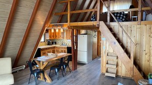 Une pièce confortable aménagée sous les toits, avec un escalier en bois, un coin repas comprenant une table et des chaises, et une kitchenette dotée de meubles en bois et d’un réfrigérateur.