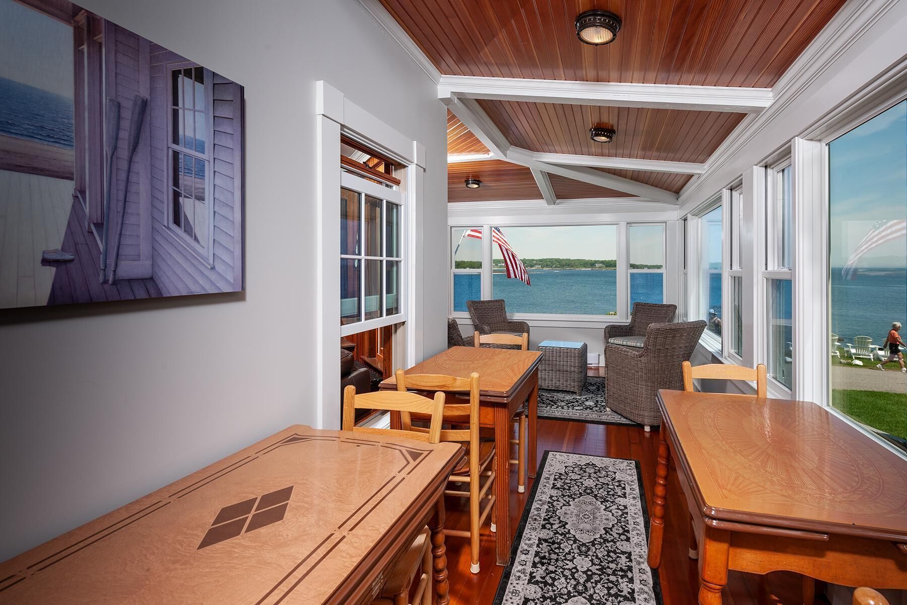 A sunroom with a view of the sea, wooden furniture, and a large painting on the wall.