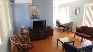A living room with a flat-screen TV on a wooden cabinet, a dining table with chairs, a wicker chair, and a sofa.