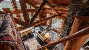 L’intérieur d’un chalet rustique, avec un plafond haut, des poutres en bois, un canapé confortable, une lampe et une vue sur l’extérieur.