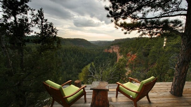 Une terrasse en bois avec deux fauteuils verts rembourrés et une petite table, donnant sur une vallée boisée.