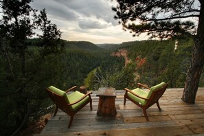A wooden deck with two green cushioned chairs and a small table, overlooking a forested valley.