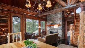 L’intérieur d’un chalet rustique, avec une table à manger en bois, une cheminée en pierre et de grandes fenêtres donnant sur une zone boisée.