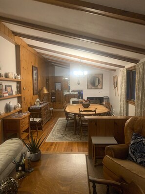 A cozy living room with a wooden dining table, a sofa, and a coffee table. There is a kitchen area in the background with wooden cabinets and a hanging light fixture. - Location! Walk to Cranmore and Town! (Conway)