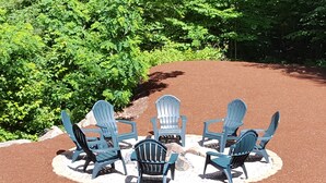 Quattro poltrone Adirondack disposte circolarmente su un'area di ghiaia, delimitate da un bordo bianco, con fitta vegetazione sullo sfondo.