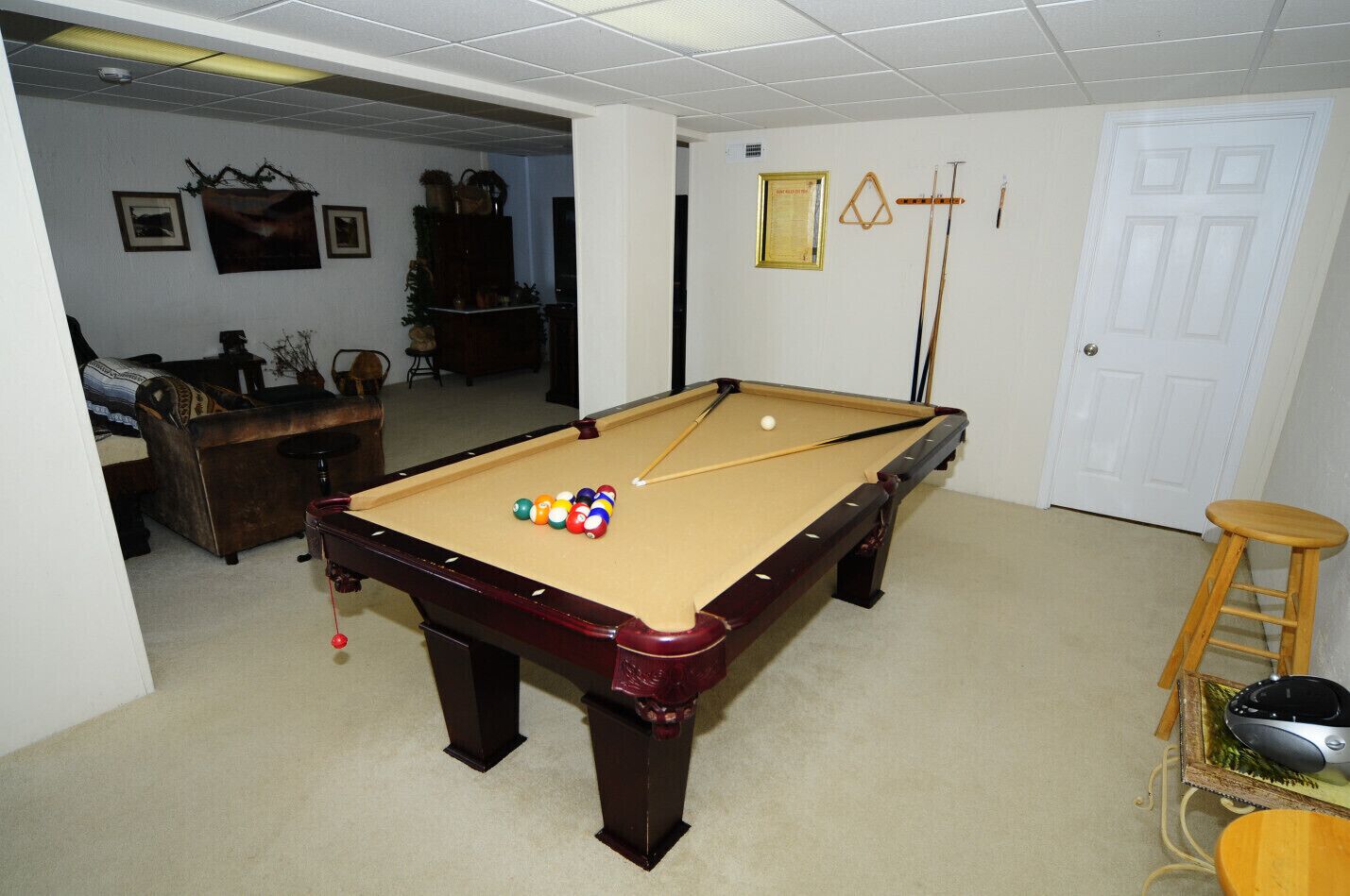 A pool table in a room with a door, a small stool, and a framed picture on the wall.