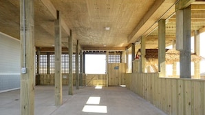 Um espaço amplo e interno, com vigas e colunas de madeira, piso de concreto e uma grande janela que permite a entrada de luz natural.