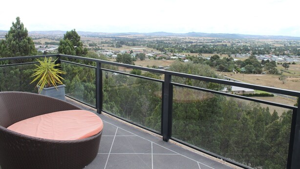 A balcony with a wicker chair and a potted plant, overlooking a town with houses and greenery. - The Winerack- spectacular views (Mudgee)