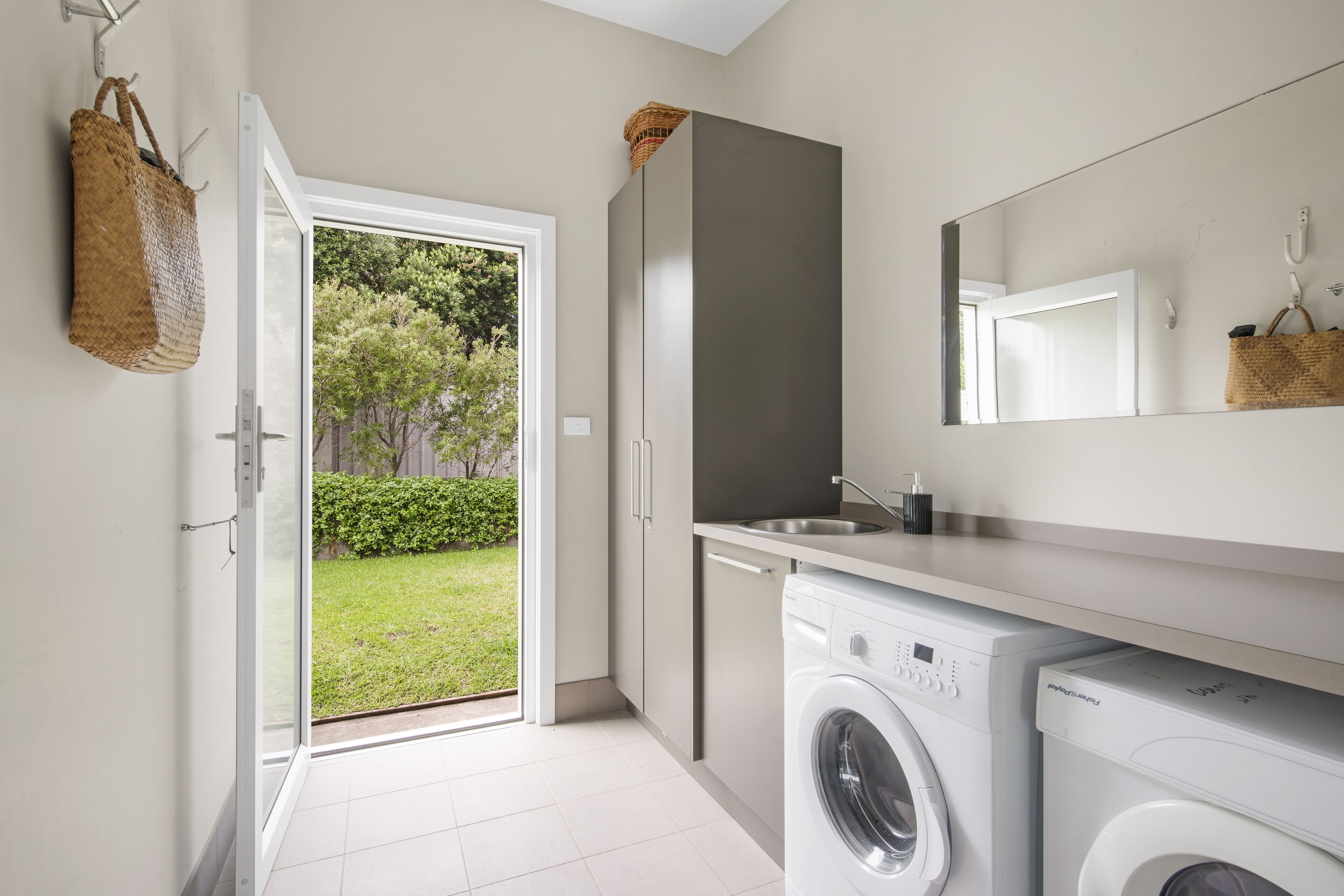 A modern laundry room with a washing machine, a dryer, and a sink. There is a basket hanging on the wall and a door leading to a garden.