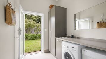 A modern laundry room with a washing machine, a dryer, and a sink. There is a basket hanging on the wall and a door leading to a garden.