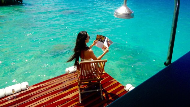 Una persona leyendo una revista en una cubierta de madera, con vista clara a un océano de agua turquesa y una estructura de techo de paja al fondo.