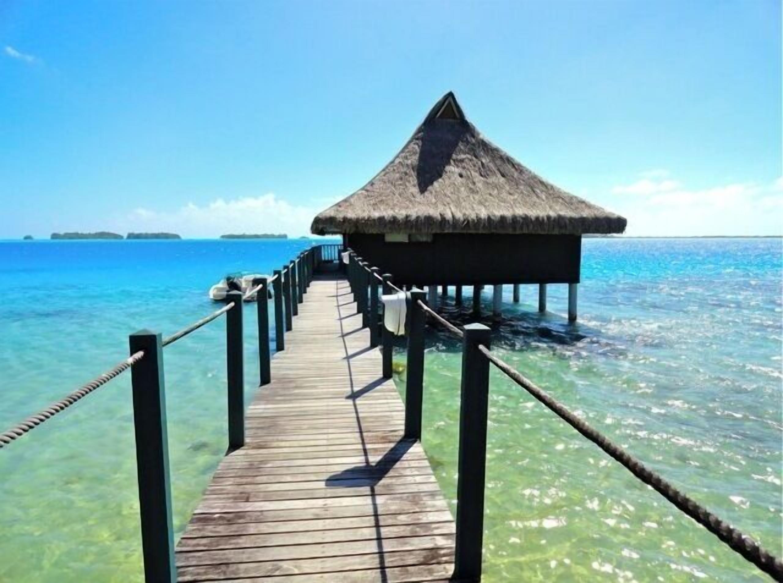 Eine Holzsteg, der zu einem über dem Wasser stehenden Bungalow mit einem Reetdach führt, mit klarem, blauem Wasser und fernen Inseln.