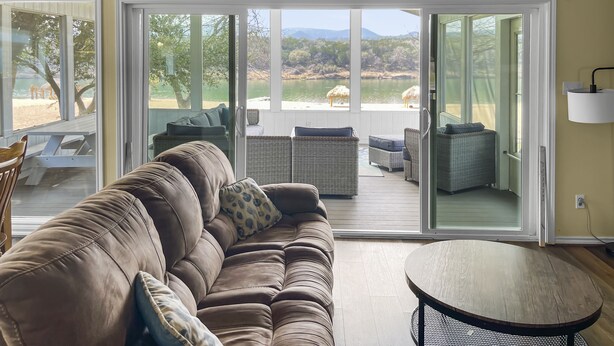 A living room with a brown leather sofa, a round wooden table, and a view of a beach through large sliding glass doors. - Waterfront, gorgeous sunsets, sandy beach, dogs allowed. (Kingsland)