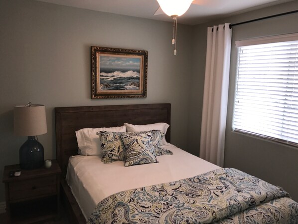 A bedroom with a bed, a nightstand, a lamp, a painting, and a window with blinds.