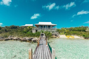 A wooden pier leading to a beachfront property with a white-roofed building and a view of the ocean.