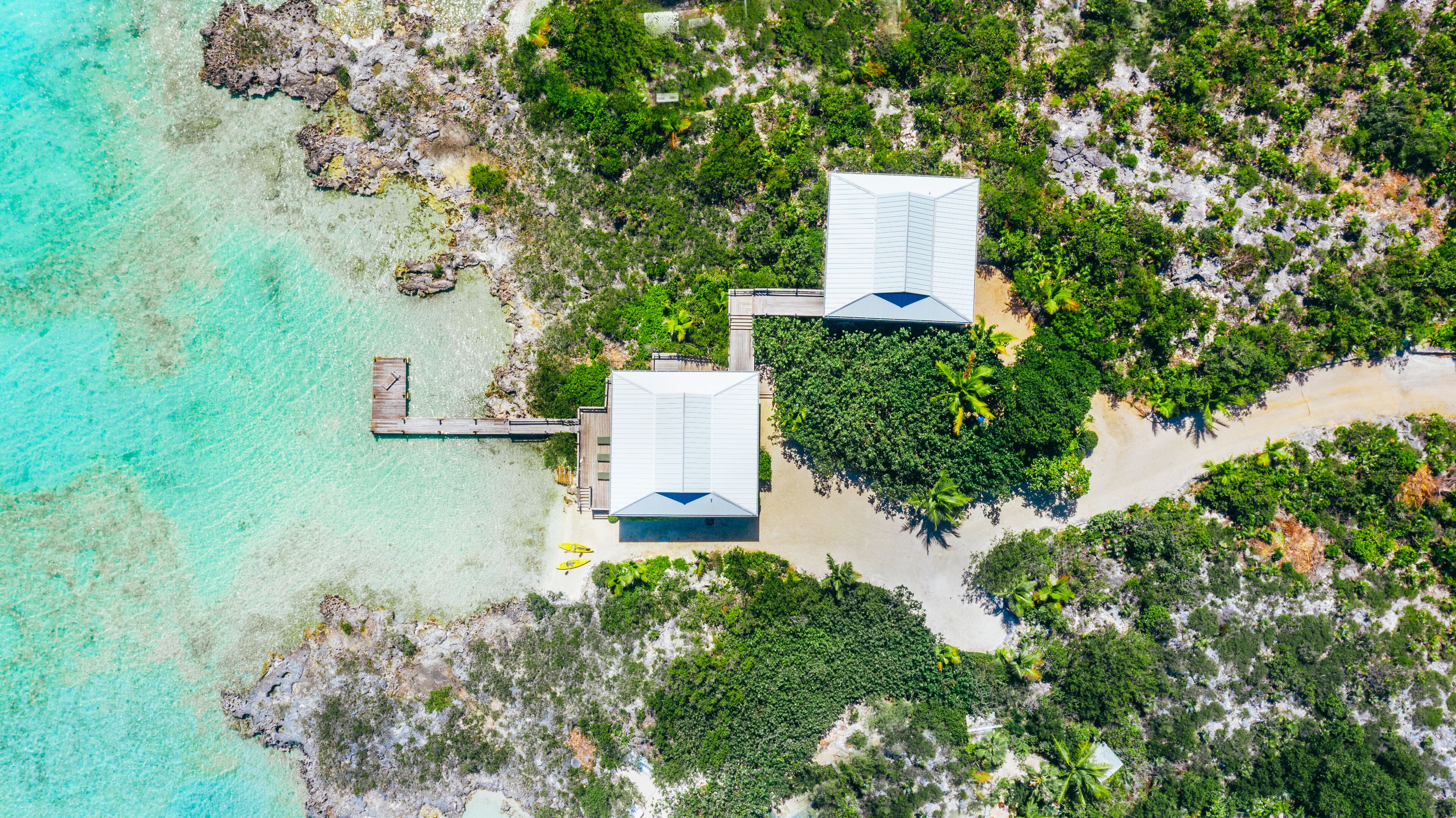 Vue aérienne d’une propriété située sur la côte, avec deux bâtiments blancs, une jetée en bois et une plage de sable bordée d’une végétation luxuriante.