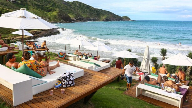 Un resort sulla spiaggia con lettini e ombrelloni, un pontile di legno e vista sull'oceano e sulle colline.