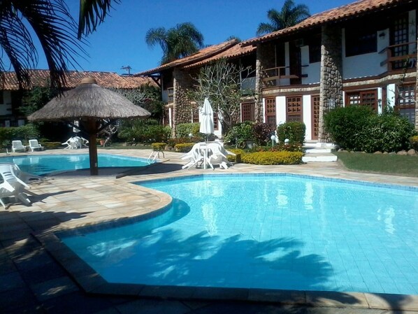 A swimming pool with lounge chairs and a thatched umbrella, surrounded by a well-maintained garden and a building with a tiled roof.