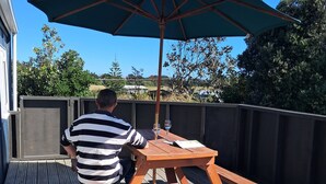 A person seated at a wooden picnic table with a large umbrella, on a wooden deck with a view of greenery and buildings in the background. - Seabury Lookout – Elevated Bach Living with Expansive Outlooks (Foxton Beach)