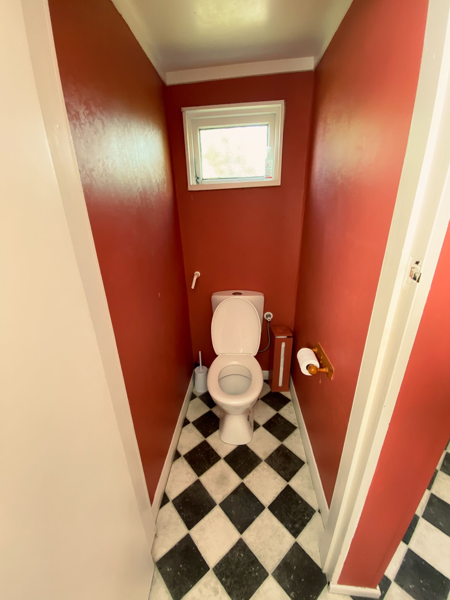 Une petite salle de bain avec des toilettes blanches, une fenêtre au-dessus des toilettes et un carrelage à motifs carrés.